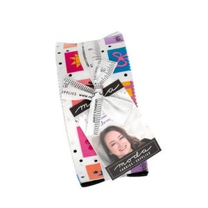 May include: A stack of fabric strips tied with a white ribbon featuring a black ruler print. The fabric has a white background with black polka dots and colorful rectangular patches containing various shapes. A tag shows a photo of a person and the text Moda.