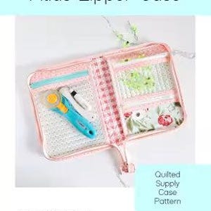 May include: Open pink quilted zipper case with multiple compartments, holding a blue and yellow rotary cutter and white pens. The case has a floral print and the text "Atlas Zipper Case".