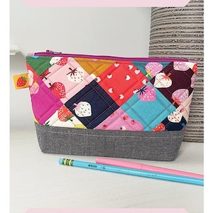 May include: A quilted fabric pouch with a patchwork design featuring strawberries and other patterns. The pouch has a gray base and a pink zipper. The text "Candy Gems Pouch" is at the top. The pouch is 6.5" x 9".