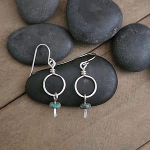 May include: A pair of silver dangle earrings featuring a circular wire frame, a small turquoise-colored bead, and a short, flat, hammered silver bar at the base. The earrings are attached to silver ear wires for hanging.