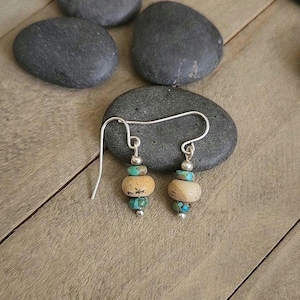 May include: Handmade earrings with a silver hook and a dangle design. Each earring features a combination of turquoise and tan-colored beads, accented with small silver beads. The earrings are displayed on a dark gray stone.