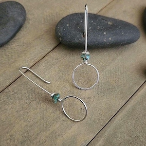 May include: A pair of silver hoop earrings with a turquoise bead accent. The earrings have a simple, elegant design, with a circular hoop and a long, curved hook. The earrings are displayed on a wooden surface.