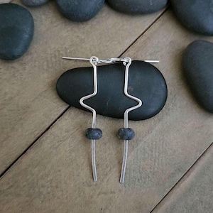 May include: A pair of silver-toned metal earrings featuring a geometric, angular wire design. Each earring consists of a single bent wire forming a sharp zigzag shape, with a small, dark grey, round bead threaded onto the lower vertical section.