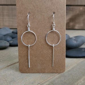 May include: A pair of silver-toned dangle earrings featuring a circular hoop design with a straight vertical bar suspended from the bottom of each circle. The earrings are attached to a rectangular brown display card with a small hole at the top.