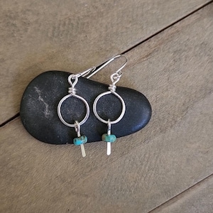 May include: Silver hoop earrings with turquoise beads and silver bar accents. The earrings have a simple, minimalist design. The earrings are displayed on a black stone against a wooden background.