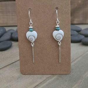 May include: A pair of dangle earrings featuring white heart-shaped beads with a grey spiral pattern, accented by small turquoise-colored beads and silver-toned metal components, including a thin vertical bar at the base of each earring.