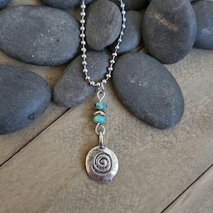 May include: A silver-toned ball chain necklace featuring a pendant with a circular charm embossed with a spiral design. The pendant is connected to the chain by two small, round, turquoise-colored beads with visible dark matrix veining.