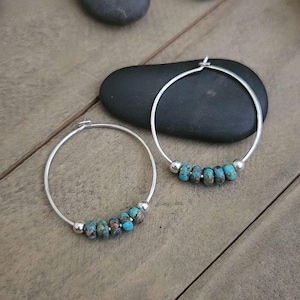 May include: A pair of silver hoop earrings featuring a row of small, round turquoise-colored beads with brown matrix patterns, accented by two smooth silver spheres on each side of the bead cluster, creating a simple and balanced jewelry design.