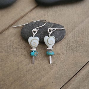 May include: A pair of dangle earrings featuring white heart-shaped shell beads with a spiral pattern, accented by small round turquoise-colored beads and silver-toned metal spacers, suspended from silver-toned metal ear wires.