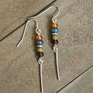 May include: A pair of dangle earrings with silver-tone hooks. Each earring features a vertical row of colorful beads in shades of orange, blue, green, and brown, with a long, thin silver bar hanging below.