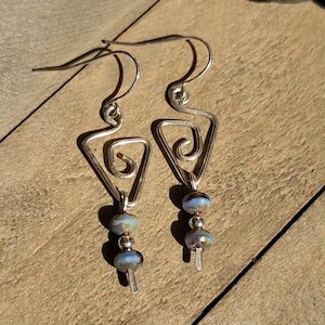 May include: A pair of silver-toned earrings with a triangular design and a spiral detail. Each earring features three blue and green glass beads. The earrings are displayed on a wooden surface.