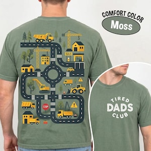 Puede incluir: Una camiseta verde musgo con un gráfico en la espalda de una obra en construcción con carreteras, camiones amarillos, grúas y edificios. El frente muestra el texto blanco "TIRED DADS CLUB" en una disposición circular con borde punteado.
