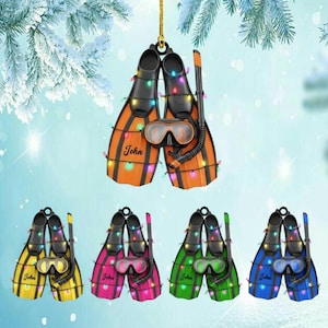 Pode incluir: Enfeite de Natal com um par de barbatanas de mergulho laranja e pretas, um snorkel e uma máscara de mergulho. O enfeite é decorado com luzes de corda e o nome "John". Outros enfeites em amarelo, rosa, verde e azul também são mostrados.