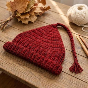 Pode incluir: Um gorro de crochê vermelho escuro feito à mão com uma longa borla trançada. O chapéu tem formato triangular e está sobre uma superfície de madeira. Uma bola de lã branca, folhas secas e agulhas de crochê também são visíveis.