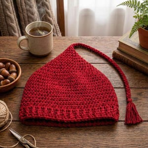 May include: A handmade, red crochet hat with a pointed top and a long, braided tassel. The hat is displayed on a wooden surface, alongside a cup of tea, acorns, and a pair of scissors. The hat's texture is visible.