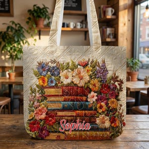 Könnte beinhalten: Cremefarbene Tragetasche mit einem Blumen- und Buchdesign. Das Design zeigt einen Stapel Bücher mit bunten Blumen und dem Namen "Sophia". Die Tasche hat cremefarbene Griffe und ein Steppmuster.