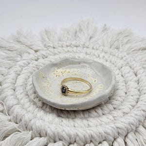 Könnte beinhalten: Ein goldener Ring mit einem kleinen, dunkelblauen ovalen Edelstein, der von einer feinen, perlenartigen Metallfassung umgeben ist. Der Ring liegt in einer kleinen, runden, cremefarbenen Keramikschale mit gesprenkelter Textur und unebenem Rand.