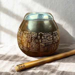 Può includere: Una tazza da mate a forma di zucca marrone con un bordo argentato e un design decorativo inciso. Una cannuccia di legno, o bombilla, è appoggiata accanto alla tazza su un panno dai toni neutri. La tazza è progettata per bere yerba mate.
