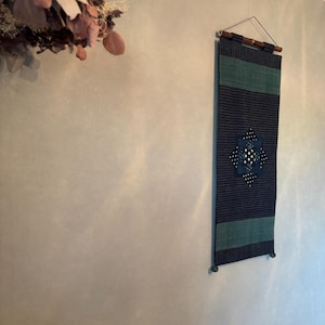 以下が含まれることがあります： 濃い青と緑のデザインの長方形の壁掛け。このテキスタイルは、白い水玉模様と幾何学模様の正方形を中央に配置しています。上部には吊り下げ用の木製の棒が付いています。