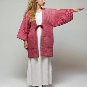 May include: A red kimono-style jacket with a textured weave and floral embroidery on the sleeves. The jacket is open in the front and tied with a dark red bow. The text "SAJU HERITAGE" is on the left side.