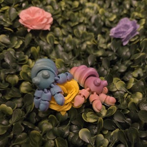 May include: Two articulated hermit crab figurines in blue and pink hues rest on a bed of yellow fabric, surrounded by green foliage. A pink and purple rose are in the background. The crab shells have a gradient color effect.
