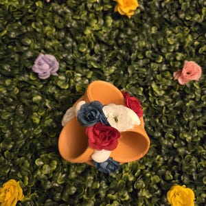 May include: Orange-colored sandals decorated with red, white, and blue fabric flowers. The sandals are set against a backdrop of green foliage and scattered artificial flowers in pink and yellow.