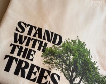 May include: Cream-colored t-shirt with the text "STAND WITH THE TREES" in bold black letters. A detailed illustration of a green tree is printed on the right side of the shirt, creating a nature-themed design.