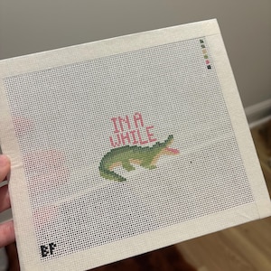 May include: A square needlepoint canvas featuring a green alligator with an open mouth and a pink tongue. Above the alligator, the text "IN A WHILE" is stitched in pink thread. The canvas has a white grid and a small black "EP" logo in the corner.