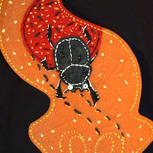 May include: Orange felt applique featuring a black beetle design. The beetle is stitched with white thread and has white eyes. The applique has a red and black dotted area and white dotted accents. The background is black.