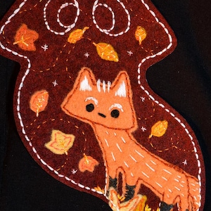 May include: A felt patch featuring an orange fox with black accents, surrounded by autumn leaves. The fox stands on a bed of flames. The patch is outlined with white stitching and set against a dark background.