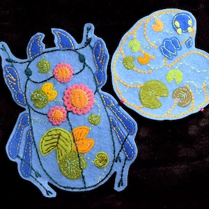 May include: Two light blue felt appliqués with embroidered details. One is a stylized insect shape, the other a lily pad design. Both feature pink, orange, green, and yellow floral and leaf accents, with gold stitching.
