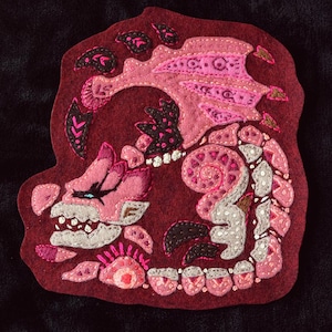 May include: A handmade felt dragon patch in shades of pink, gray, and black, with intricate embroidered details. The dragon is set against a burgundy background. The patch features a detailed design of a dragon with wings and a curled tail.