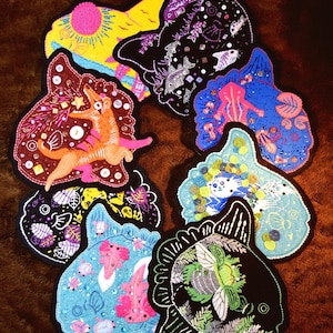 May include: A collection of seven distinct embroidered patches featuring varied designs including a yellow sun, a black fish, a pink axolotl, a green beetle, and an orange creature, all detailed with colorful thread, sequins, and small buttons.