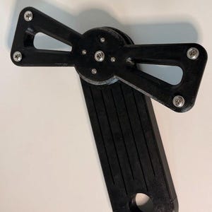 May include: A black mechanical component featuring multiple arms and a central pivot, held together with silver screws. The component has a rectangular base with a circular hole and parallel grooves.