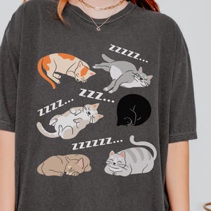 May include: Dark gray t-shirt featuring illustrations of sleeping cats in various poses, with "Zzz..." text above them. The cats are in shades of orange, gray, brown, and black. A casual, comfortable tee for cat lovers.