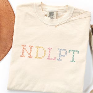 Op de afbeelding: Een crèmekleurig t-shirt met korte mouwen en de tekst NDLPT geborduurd in een veelkleurig kruissteekpatroon. De letters zijn oranje, geel, roze, blauw en groen. Het t-shirt heeft een Comfort Colors-merklabel bij de binnenhalslijn.