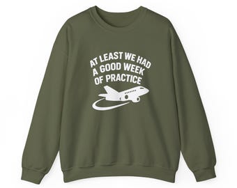 May include: An olive green sweatshirt featuring the text "AT LEAST WE HAD A GOOD WEEK OF PRACTICE" and a white airplane graphic. The airplane has a curved trail. The sweatshirt is a casual style.