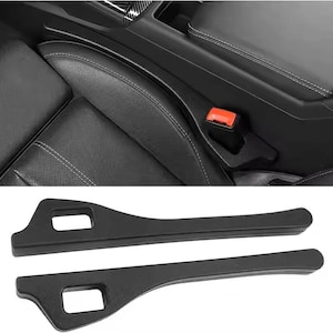 May include: Black car interior with leather seats and a seat belt buckle. Two black plastic car interior trim pieces are in the foreground, each with a rectangular cutout.