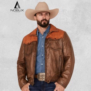 Op de afbeelding: Bruine leren jas met een contrasterende oranje pas en zakken. De jas heeft een klassieke westernstijl met een knoopsluiting en een decoratieve gesp. Gedragen met een spijkeroverhemd en een cowboyhoed.