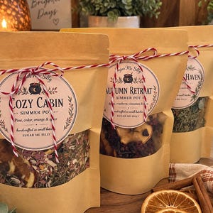 May include: Three stand-up kraft paper pouches containing dried botanical simmer pot ingredients. Each bag features a round label with the text Sugar Me Silly, Cozy Cabin, or Autumn Retreat, tied with red and white baker's twine at the top.