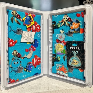 May include: A display case featuring a blue fabric with cartoon characters from the movie "Finding Nemo." The case holds various pins, including Nemo, Dory, and other characters. Some pins are in packaging, and the case is open.