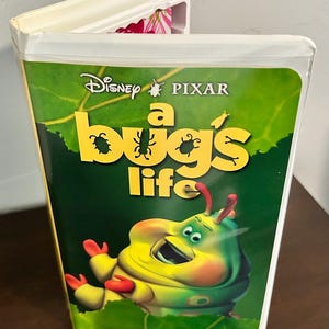 Può includere: Cassetta VHS di A Bug's Life. La copertina presenta i loghi Disney e Pixar, il titolo in giallo e un'immagine a cartoni animati di un bruco verde. Lo sfondo è una foglia verde.
