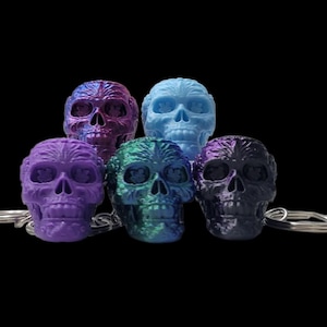 May include: Five colorful skull keychains in purple, blue, green, and black hues. Each skull has detailed facial features and is attached to a silver keyring. The keychains are set against a black background.