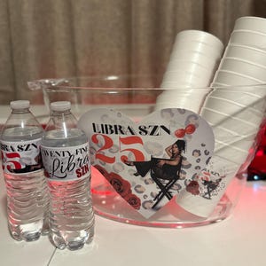 May include: A clear plastic ice bucket filled with white paper cups and two water bottles. The water bottles have labels that read "Libra SZN 25". A heart-shaped sign inside the bucket reads "Libra SZN 25".