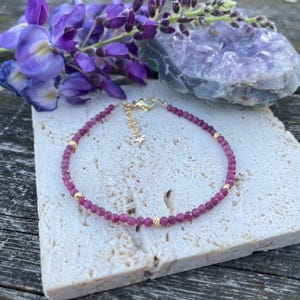 Könnte beinhalten: Ein zartes Armband mit kleinen, facettierten magentafarbenen Perlen, die mit winzigen, runden goldfarbenen Metallzwischenstücken durchsetzt sind. Das Armband hat eine goldfarbene Verlängerungskette mit einem kleinen sternförmigen Anhänger.
