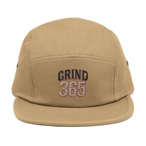 May include: A tan five-panel cap with a flat peak. The cap displays the text "GRIND 365" in a black and silver design on the front. The cap is made of a durable fabric.