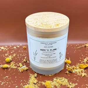May include: A frosted glass candle with a wooden lid. The label reads "HOPE'S FLAME" with "Lavender & Chamomile" and "Natural Candle 7 oz (198g)". The candle is surrounded by dried flowers on a brown surface.