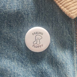 May include: A white button badge with the words "FEELING FLOPPY" and a cartoon blob illustration. The badge is on a blue denim jacket with a beige corduroy collar.