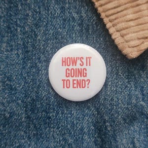 May include: A white button badge with the red text "HOW'S IT GOING TO END?". The badge is pinned to a blue denim jacket, with a tan corduroy collar in the corner.