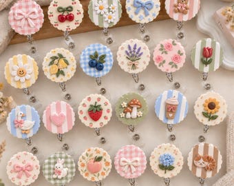 May include: A collection of twenty-four circular badge reels featuring scalloped edges and various decorative motifs including bows, fruits, flowers, animals, and coffee cups, each attached to a retractable cord mechanism with a metal clip.